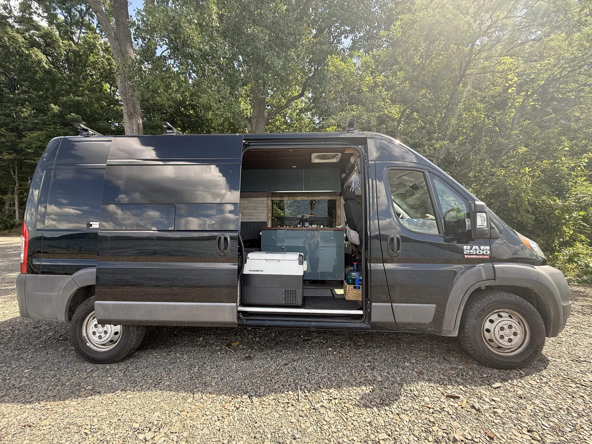 Our black 2017 Ram ProMaster 2500 high roof parked on gravel with the sliding door open, showing the galley cabinets and the chest fridge inside