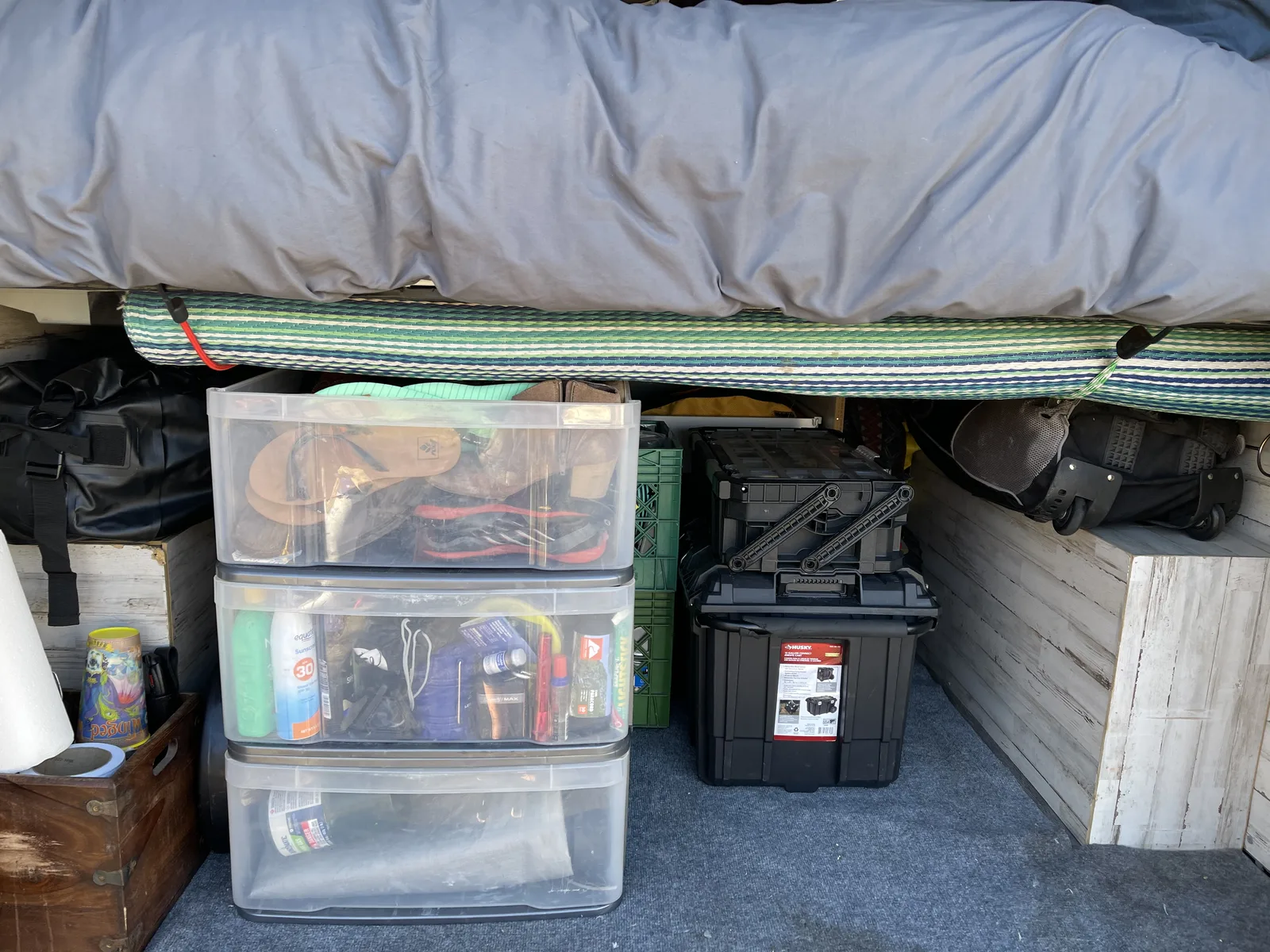 The under-bed garage seen through the open back doors: a three-drawer plastic storage container, a hard toolbox, a rolled camp rug, duffel bags and hiking boots packed under the mattress