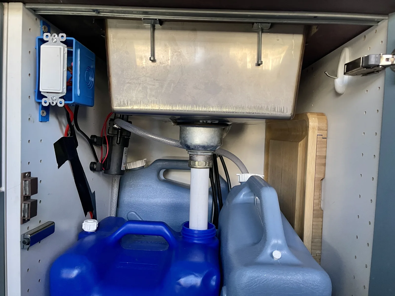 Under the sink in our van: the stainless basin, the water pump, and the fresh and grey water jugs stood upright beneath it